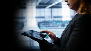 Businesswoman in a dark blazer using a tablet in a modern office; blurred background shows screens and daylight.