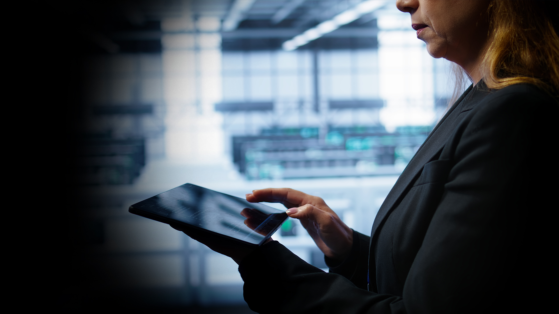 Businesswoman in a dark blazer using a tablet in a modern office; blurred background shows screens and daylight.
