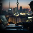 Person typing on a laptop at a dimly lit outdoor table with a blurred city skyline at dusk behind them.