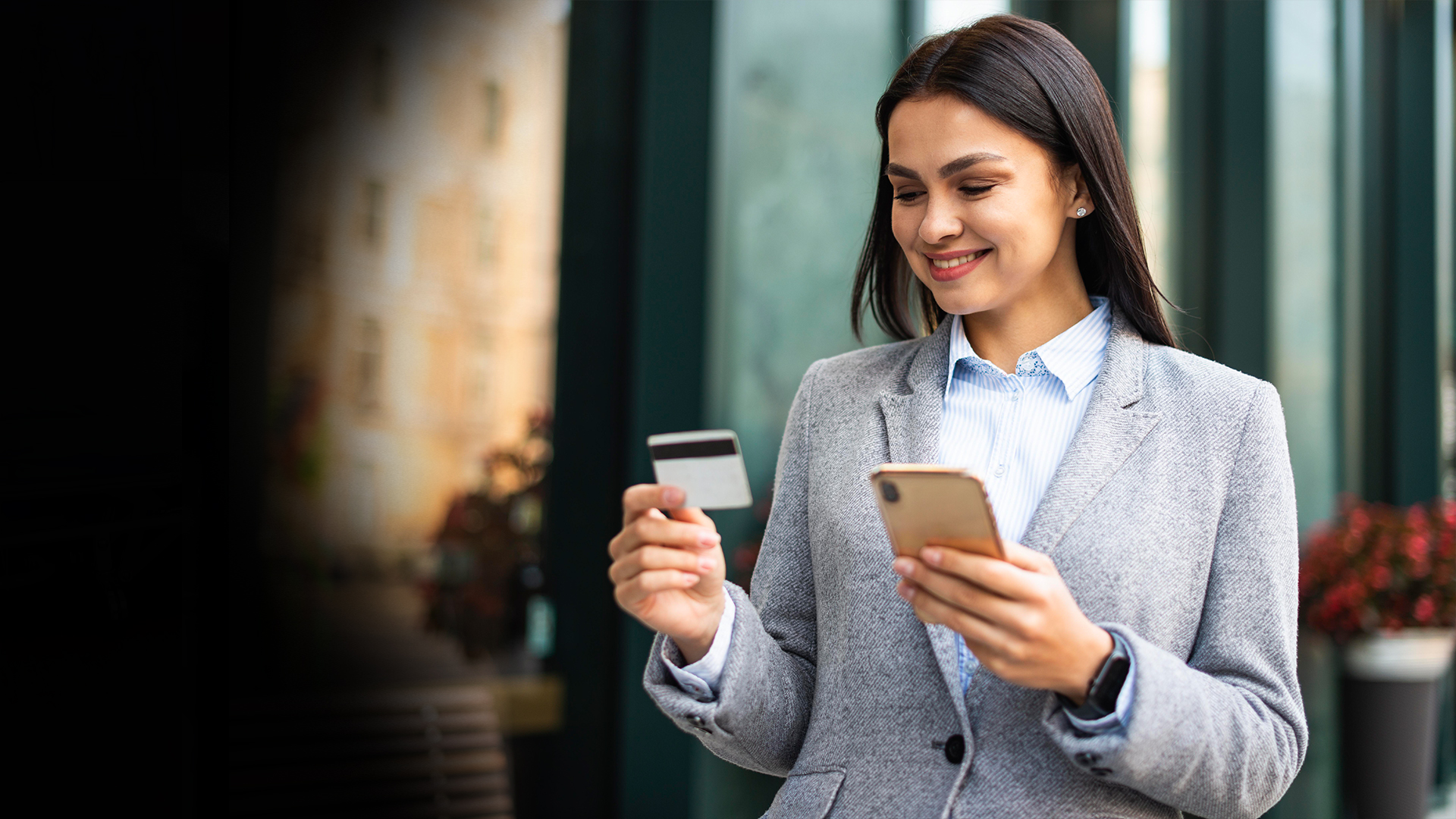 Businesswoman in a gray blazer smiling outdoors while holding a smartphone and a credit card, ready to pay.